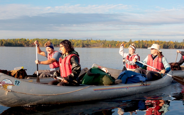 A group of students wearing life jackets smile as they paddle canoes on calm water. In the distance, trees line the shore. 